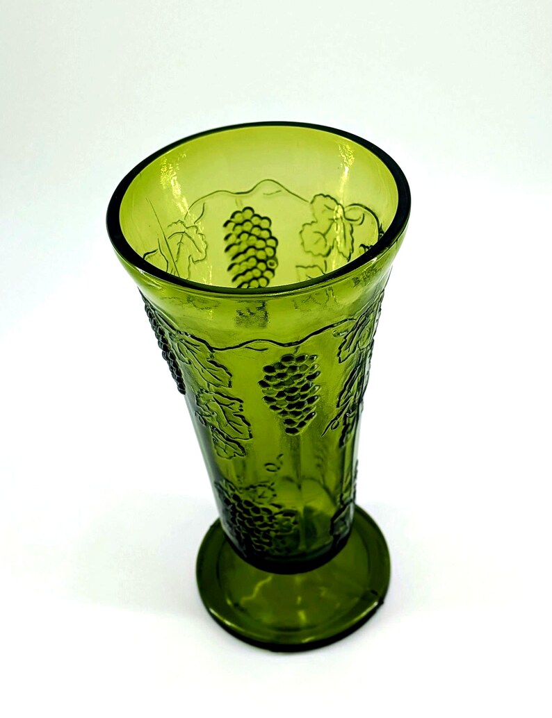 Op de afbeelding: Een groene glazen vaas met een uitlopende bovenkant en een ronde basis. De vaas is versierd met reli&euml;f druiven- en bladerenontwerpen. Het glas heeft een lichte groene tint en de vaas is waarschijnlijk vintage.