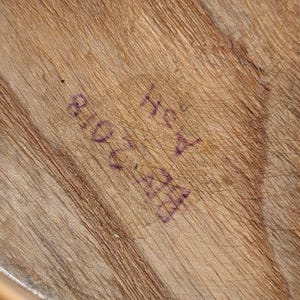 K&ouml;nnte beinhalten: Nahaufnahme einer Holzoberfl&auml;che mit sichtbarer Maserung und Textur. Die Oberfl&auml;che hat eine hellbraune Farbe mit dunkleren braunen Streifen. Auf dem Holz ist Text mit violetter Tinte gestempelt, einschlie&szlig;lich der Zahlen "810 2028" und der Buchstaben "Hey A".