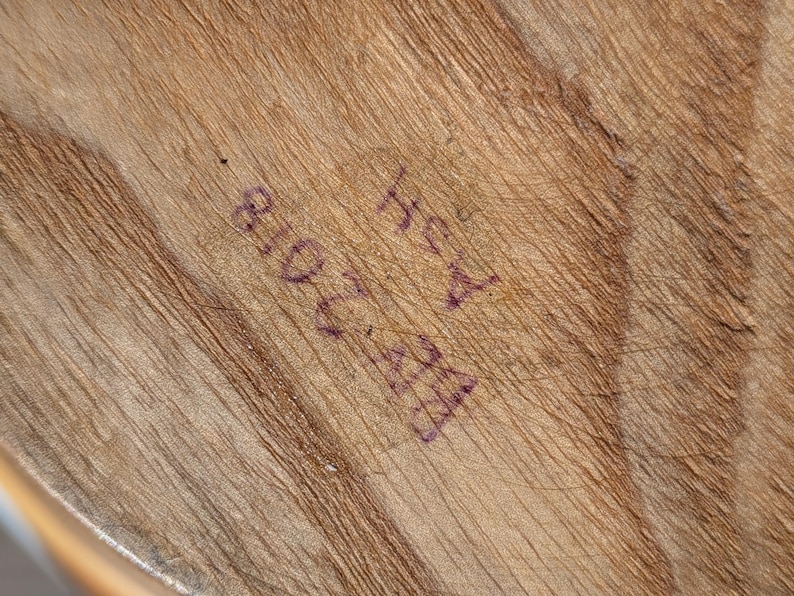 K&ouml;nnte beinhalten: Nahaufnahme einer Holzoberfl&auml;che mit sichtbarer Maserung und Textur. Die Oberfl&auml;che hat eine hellbraune Farbe mit dunkleren braunen Streifen. Auf dem Holz ist Text mit violetter Tinte gestempelt, einschlie&szlig;lich der Zahlen "810 2028" und der Buchstaben "Hey A".