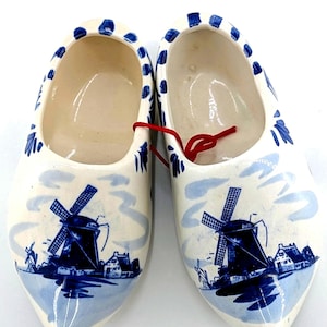 May include: Pair of white ceramic clogs with a blue windmill and landscape design. The clogs have a rounded toe and are decorated with blue floral accents around the top edge. A red string is tied between the clogs.