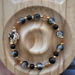 Pode incluir: Uma pulseira de contas pretas, transparentes e laranja, com detalhes dourados. Inclui um pequeno pingente de tartaruga com manchas pretas e brancas. A pulseira é exibida em uma bandeja de madeira em forma de gato.