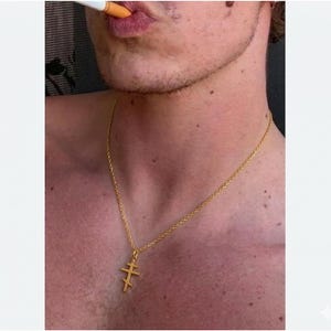 May include: A gold-colored necklace with a double-barred cross pendant hangs around a person's neck. The chain is delicate, and the cross has three horizontal bars. The person is smoking a cigarette.