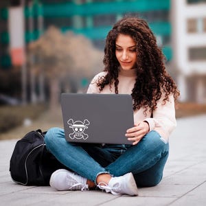 May include: A person sitting cross-legged, using a gray laptop with a white Jolly Roger sticker. The person is wearing a light pink sweater, blue jeans, and white sneakers. A black backpack is next to them.