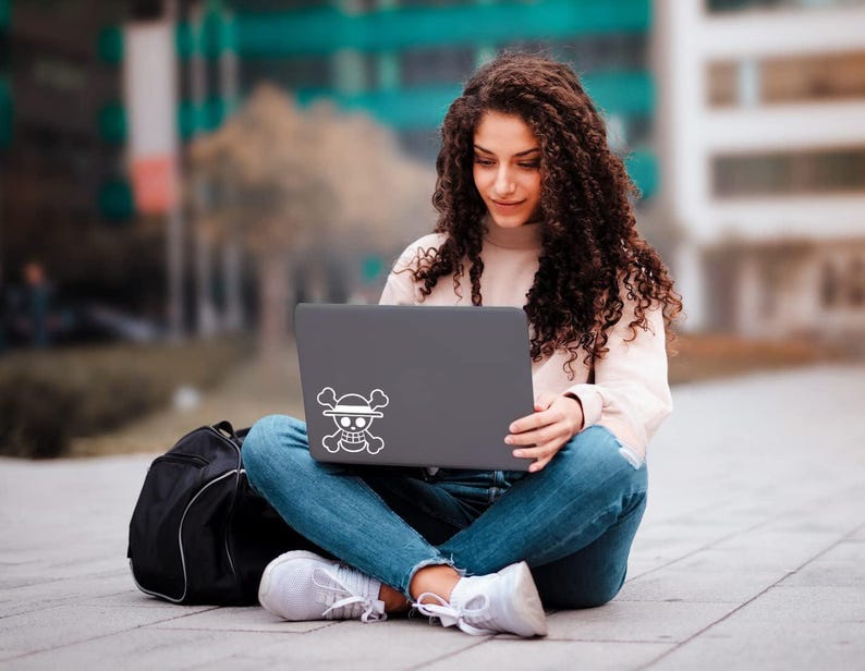 May include: A person sitting cross-legged, using a gray laptop with a white Jolly Roger sticker. The person is wearing a light pink sweater, blue jeans, and white sneakers. A black backpack is next to them.
