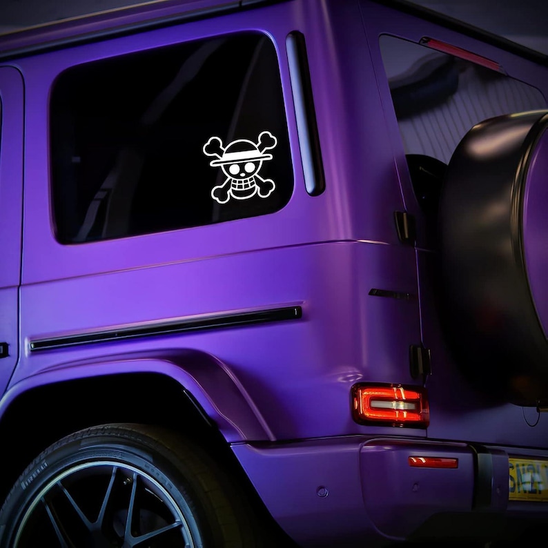 May include: A white One Piece Jolly Roger skull and crossbones sticker is affixed to the rear window of a purple SUV. The sticker features a skull wearing a straw hat, with crossbones beneath.