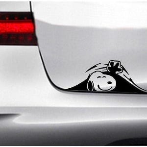 May include: A black and white Snoopy car decal. The decal shows Snoopy peeking out from behind a car's trunk, waving with a smiling expression. The decal is applied to the white trunk of a car, with the taillight visible.