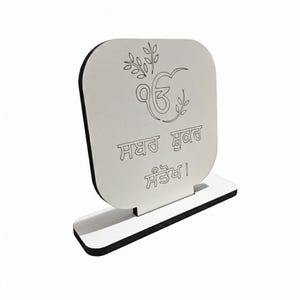 May include: A white wooden decorative plaque with a black border and base. The plaque features engraved text and a symbol with leaf accents. The text is in a stylized script. The plaque is designed to stand upright.