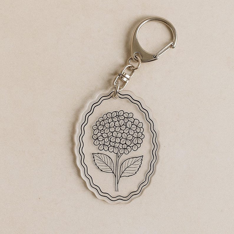 May include: Clear acrylic keychain with a silver-colored clasp. The keychain features a black and white illustration of a hydrangea flower within an oval frame. The flower has detailed petals and leaves.