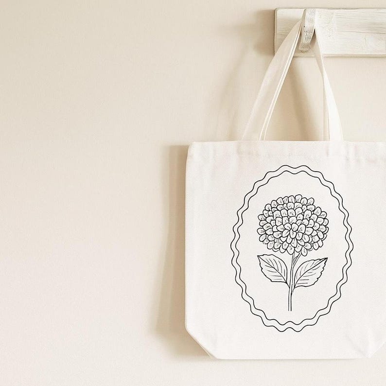 May include: White tote bag with a black and white floral design. The tote bag features a detailed illustration of a hydrangea flower within a decorative oval frame. The bag has two white handles and is hanging on a wooden hook.