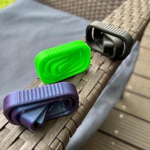 Könnte beinhalten: Drei kleine, farbenfrohe 3D-gedruckte Objekte. Eines ist leuchtend grün, ein anderes schimmert lila und blau, und das dritte ist schwarz. Alle haben eine ähnliche rechteckige Form mit einem gewölbten Innenraum. Sie liegen auf einer braunen, gewebten Oberfläche.