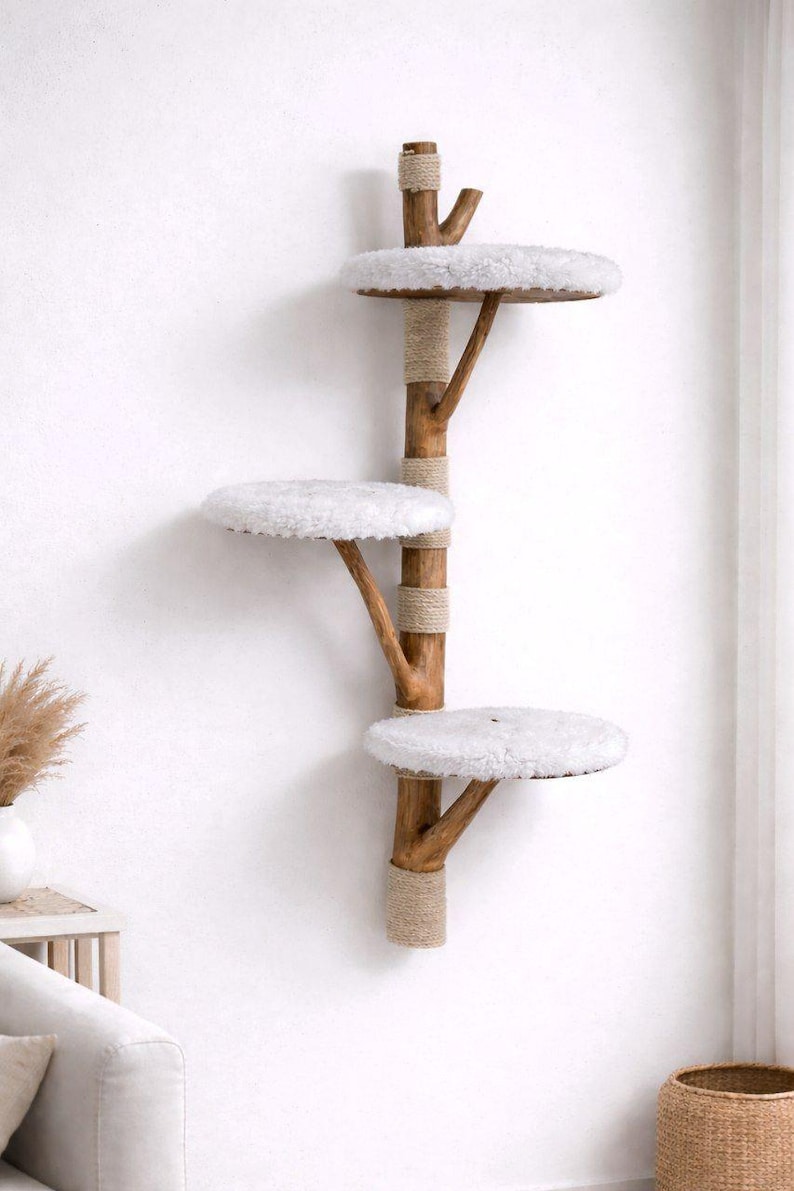 May include: A wall-mounted cat tree with three circular platforms covered in white, fluffy material. The platforms are supported by wooden branches attached to a central wooden pole wrapped with natural rope. The cat tree is designed to be mounted on a white wall.
