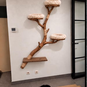 May include: A wall-mounted cat tree constructed from a natural wooden branch with a dark brown finish. It features three circular platforms topped with plush, cream-colored cushions and one rectangular wooden shelf. Sections of the branch are wrapped in light tan rope.