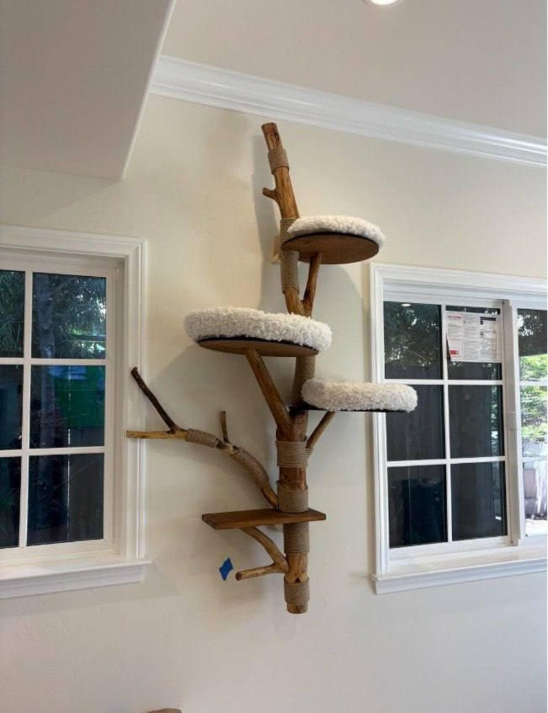May include: A wall-mounted cat tree constructed from a natural wooden branch with multiple levels. It features three circular platforms topped with white plush cushions and one lower rectangular wooden shelf, all secured with tan rope accents.