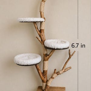 May include: A wall-mounted cat tree featuring a natural wood branch structure with three circular platforms topped with white plush cushions. The branch is wrapped with tan rope at various joints. A small section measures 17.02 cm in height.