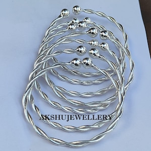 May include: A collection of silver-colored, twisted wire bracelets. Each bracelet features a small, round ball at the ends. The bracelets are arranged in a circular pattern, with the text "AKSHUJEWELLERY" visible at the bottom.