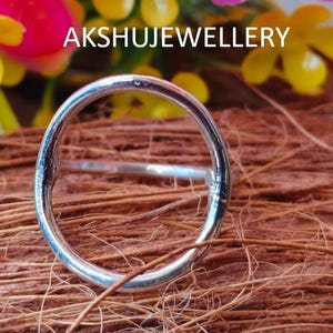 May include: A silver ring with a smooth, polished surface. The ring is set against a backdrop of colorful flowers and brown fibers. The text "AKSHUJEWELLERY" is visible.