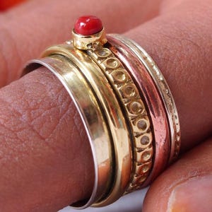 May include: A collection of rings in gold, silver, and copper tones. The rings are stacked on a finger, with a small red gemstone on the top ring. The rings have various textures and designs.