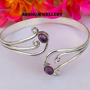 May include: Open silver cuff bracelet with two purple gemstones. The bracelet has a decorative swirl design and the text "AKSHUJEWELLERY". The bracelet is displayed against a background of pink and yellow flowers.