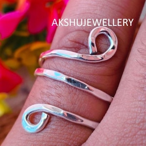 May include: A silver-colored ring wraps around a finger, featuring a spiral design with curved ends. The ring has a polished finish and is set against a background of skin and pink flowers. The text "AKSHUJEWELLERY" is visible.