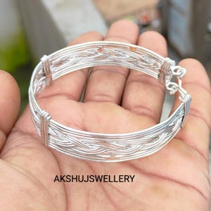 May include: A silver-colored bracelet with a woven design. The bracelet is made of thin, intertwined wires, creating a textured surface. The clasp is visible on the right side. The text "AKSHUJSJEWELLERY" is at the bottom.
