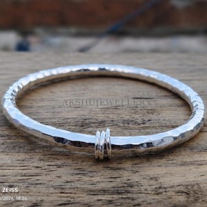 May include: A silver hammered bangle bracelet with three small rings attached. The bracelet has a textured surface and is displayed on a wooden surface. The brand name "AKSHUJEWELLERY" is visible.