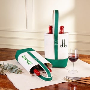 Puede incluir: Dos portadores de botellas de vino de lona, uno en posición vertical y otro de lado, ambos con detalles en blanco y verde. El portador vertical tiene un asa verde y un monograma. El otro portador tiene el nombre "Lindsey" impreso. Ambos portadores contienen botellas de vino tinto.