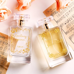 May include: Two perfume bottles with gold floral designs and clear crystal stoppers. One bottle reads "Courtney Lauren 7.28.21" and the other reads "Jovan Musk". The bottles are on a white surface with sheet music and peach roses.