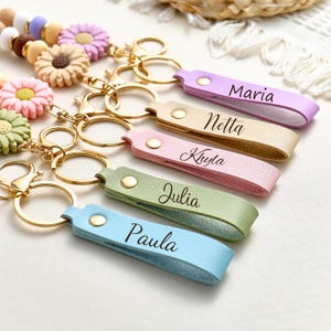 May include: A collection of personalized keychains with leather straps in various pastel colors, each engraved with a name. The keychains feature gold-colored hardware and are adorned with decorative flower charms and beads.