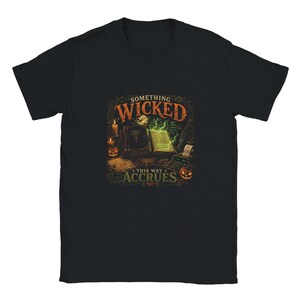 Può includere: Una t-shirt nera a maniche corte con una stampa grafica di un libro aperto con pagine verde brillante, una calcolatrice e zucche. Il testo SOMETHING WICKED THIS WAY ACCRUES è scritto in lettere arancioni e verdi con decorazioni floreali.