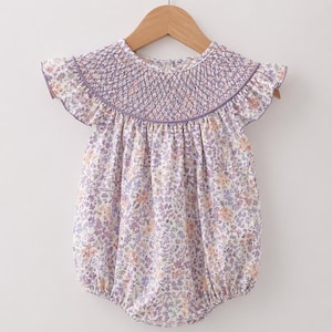 May include: A floral print baby romper with a smocked bodice and ruffled sleeves. The romper features a pattern of small purple, pink, and orange flowers on a white background. The neckline and sleeves are trimmed with a lavender border. The garment is hanging on a wooden hanger.