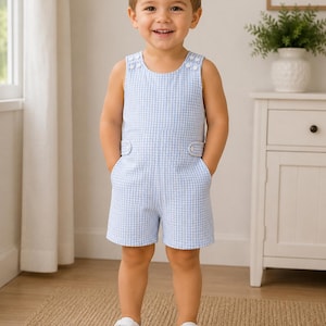 May include: A young child wearing a light blue and white gingham romper with white sneakers. The romper has decorative buttons on the shoulders and pockets on the shorts. The child is smiling and standing on a woven rug.