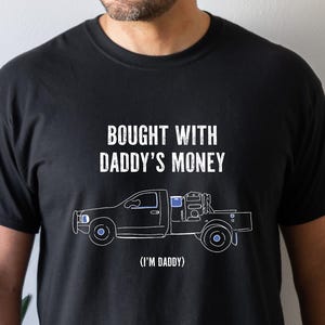 May include: Black t-shirt with the text "BOUGHT WITH DADDY'S MONEY" above a line drawing of a pickup truck. Below the truck is the text "(I'M DADDY)". The shirt is a crew neck style.