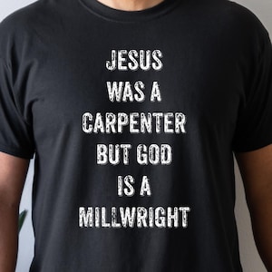 Op de afbeelding: Zwart T-shirt met de witte tekst "JESUS WAS A CARPENTER BUT GOD IS A MILLWRIGHT". Het shirt heeft een ronde hals en is gemaakt van een zacht materiaal. Het shirt wordt door een persoon gedragen.