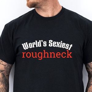 Può includere: T-shirt nero con la scritta "World's Sexiest roughneck" in bianco e rosso. La maglietta è a girocollo e viene indossata da una persona.