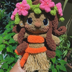 K&ouml;nnte beinhalten: Handgefertigte H&auml;kelpuppe mit braunem Teint, die einen Blumenkranz mit rosa Bl&uuml;ten und gr&uuml;nen Bl&auml;ttern tr&auml;gt. Die Puppe hat gro&szlig;e schwarze Augen, braune Locken und ein orangefarbenes und beigefarbenes Outfit. Die Puppe h&auml;lt einen beigefarbenen Rock.