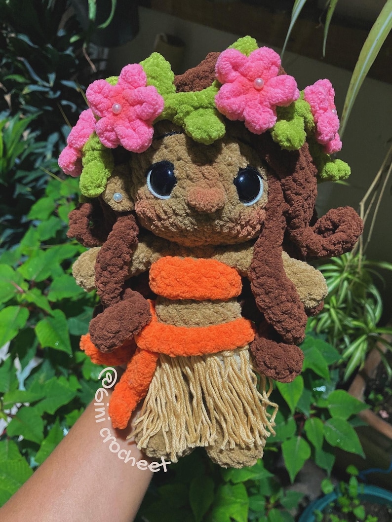 K&ouml;nnte beinhalten: Handgefertigte H&auml;kelpuppe mit braunem Teint, die einen Blumenkranz mit rosa Bl&uuml;ten und gr&uuml;nen Bl&auml;ttern tr&auml;gt. Die Puppe hat gro&szlig;e schwarze Augen, braune Locken und ein orangefarbenes und beigefarbenes Outfit. Die Puppe h&auml;lt einen beigefarbenen Rock.