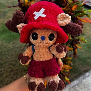 May include: A handmade crochet plush toy of a cartoon character. The toy has a red hat with a white cross, brown antlers, and red shorts. The plush is tan with black eyes and a blue nose. The toy is approximately 25 cm tall.
