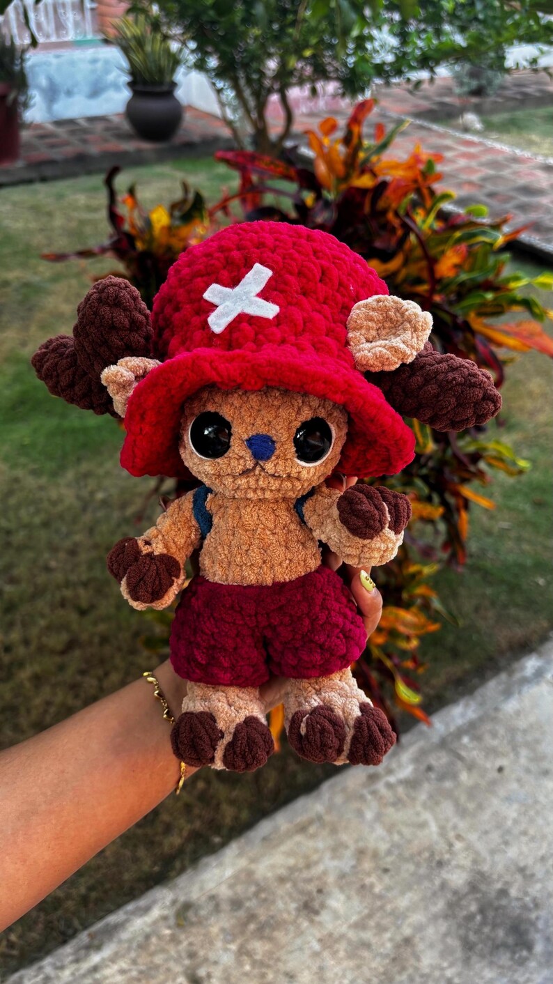 Op de afbeelding: Een handgemaakt gehaakt pluche speelgoed van een stripfiguur. Het speelgoed heeft een rode hoed met een wit kruis, bruine geweien en een rode korte broek. Het pluche is bruin met zwarte ogen en een blauwe neus. Het speelgoed is ongeveer 25 cm hoog.