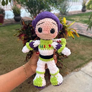 Puede incluir: Muñeca de ganchillo hecha a mano de un personaje de dibujos animados, con un casco morado, un traje verde y blanco y botas blancas con detalles verdes. La muñeca tiene ojos negros y una nariz rosa. La muñeca se sostiene frente a un fondo verde.