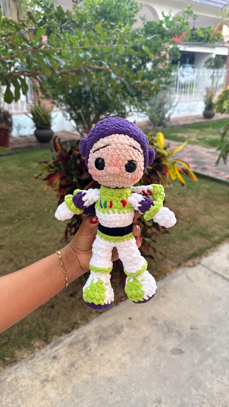 May include: A handmade crochet doll of a cartoon character, with a purple helmet, a green and white suit, and white boots with green accents. The doll has black eyes and a pink nose. The doll is held in front of a green background.