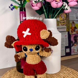 Op de afbeelding: Een handgemaakt gehaakt pluche speelgoed van een personage met een rode hoed en korte broek, een wit kruis op de hoed en bruine vacht. Het speelgoed wordt gepresenteerd op een geweven mat, met Lego-bloemen op de achtergrond.