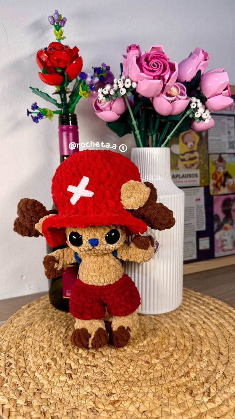 Op de afbeelding: Een handgemaakt gehaakt pluche speelgoed van een personage met een rode hoed en korte broek, een wit kruis op de hoed en bruine vacht. Het speelgoed wordt gepresenteerd op een geweven mat, met Lego-bloemen op de achtergrond.