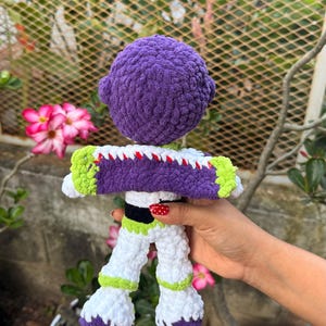 May include: A handmade crochet doll resembling a space ranger, with a purple head, white and purple suit, and green detailing. The doll is held against a backdrop of pink flowers and foliage.