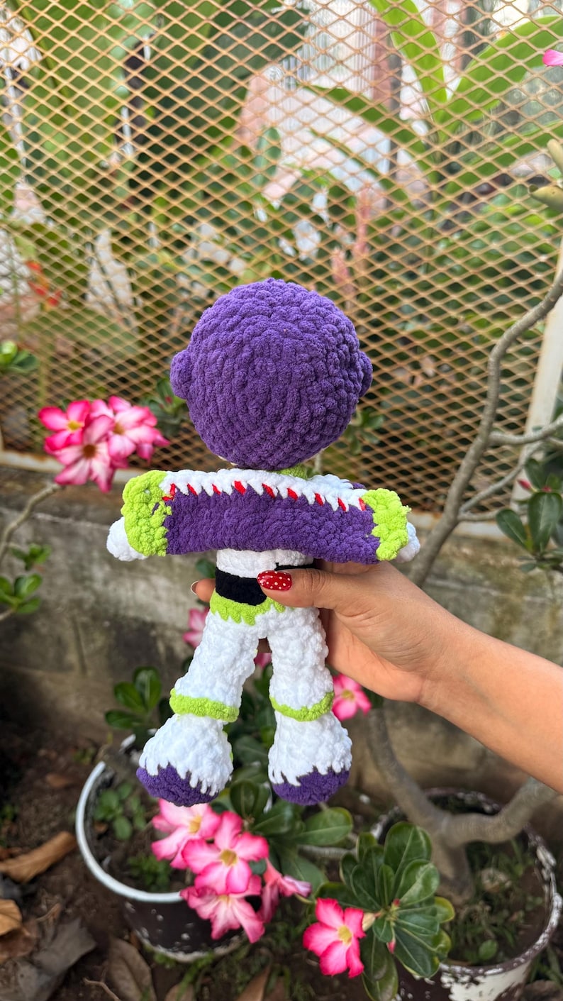 May include: A handmade crochet doll resembling a space ranger, with a purple head, white and purple suit, and green detailing. The doll is held against a backdrop of pink flowers and foliage.