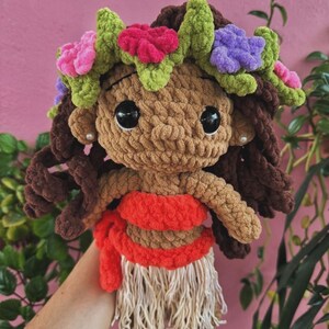 K&ouml;nnte beinhalten: Handgefertigte H&auml;kelpuppe mit brauner Haut, dunkelbraunem Haar und gro&szlig;en schwarzen Augen. Die Puppe tr&auml;gt einen roten und beigefarbenen Rock, ein rotes Oberteil und einen Blumenkranz mit rosa, lila und gr&uuml;nen Akzenten.