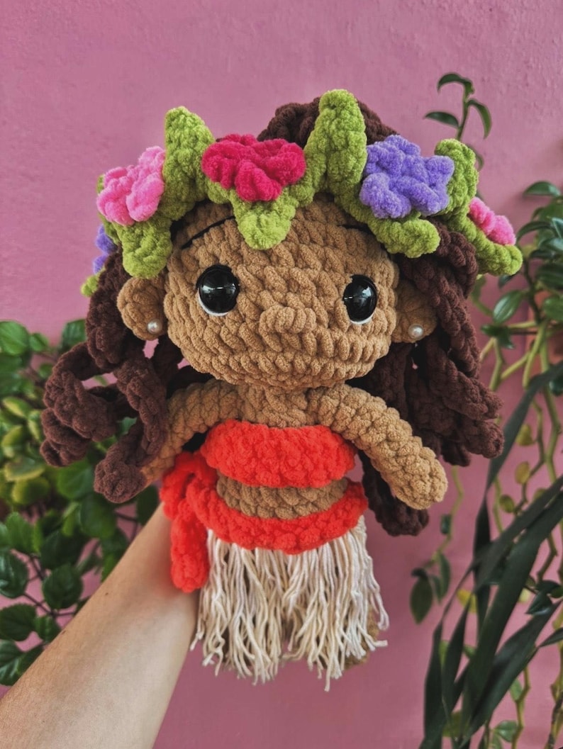 K&ouml;nnte beinhalten: Handgefertigte H&auml;kelpuppe mit brauner Haut, dunkelbraunem Haar und gro&szlig;en schwarzen Augen. Die Puppe tr&auml;gt einen roten und beigefarbenen Rock, ein rotes Oberteil und einen Blumenkranz mit rosa, lila und gr&uuml;nen Akzenten.