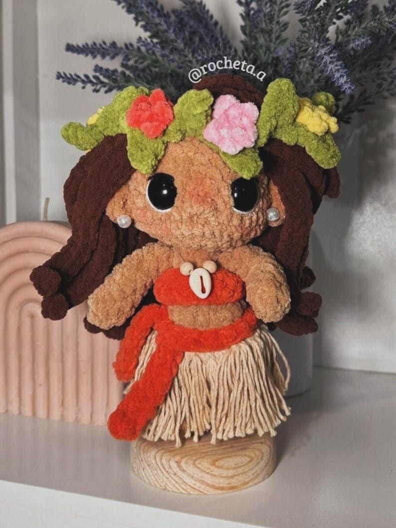 K&ouml;nnte beinhalten: Handgefertigte Pl&uuml;schpuppe mit braunen Haaren, gro&szlig;en schwarzen Augen und einem Blumenkranz. Die Puppe tr&auml;gt ein rotes Oberteil und einen Rock und steht auf einem Holzsockel. Das Gesicht der Puppe zeigt ein L&auml;cheln und Perlenohrringe.