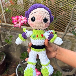 May include: A handmade crochet doll of a cartoon character, with a purple helmet, white and green spacesuit, and black accents. The doll is held in front of a blurred background of plants and flowers. The doll's height is approximately 20 cm.