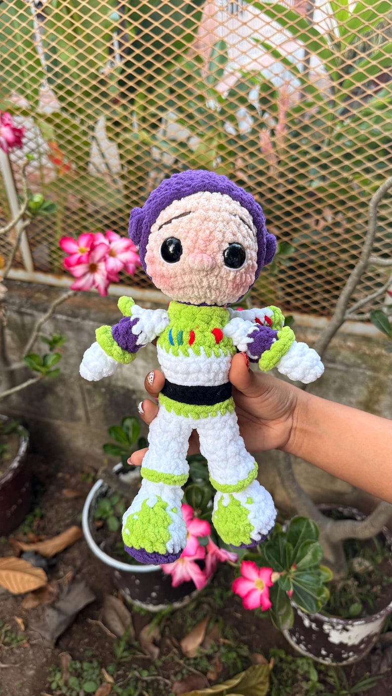 May include: A handmade crochet doll of a cartoon character, with a purple helmet, white and green spacesuit, and black accents. The doll is held in front of a blurred background of plants and flowers. The doll's height is approximately 20 cm.