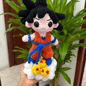 Puede incluir: Figura de ganchillo de un personaje con pelo negro puntiagudo, que lleva un traje naranja con detalles azules. La figura está sobre una nube blanca con bolas amarillas y rojas en forma de estrella. Hecho a mano.